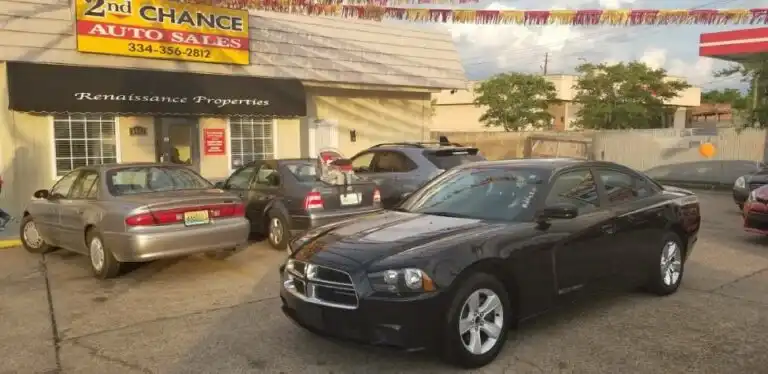 Black Dodge Charger parked at a used-car lot with a “2nd Chance Auto Sales” sign and a Renaissance Properties storefront in the background.